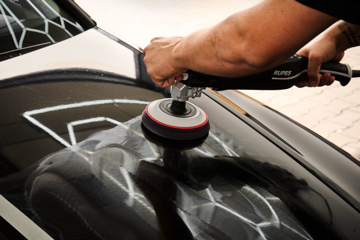 Hände polieren eine schwarze Autokarosserie mit einer Poliermaschine.